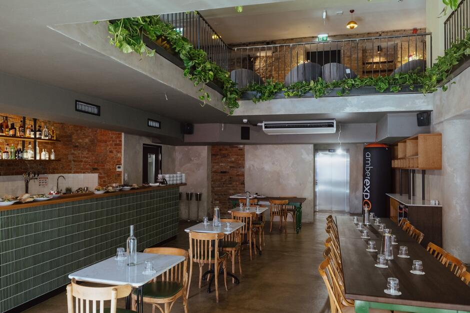 Na zdjęciu widoczna jest nowoczesna restauracja z antresolą, ozdobioną roślinami zwisającymi z balustrady. Wnętrze charakteryzuje się industrialnym stylem z odkrytymi cegłami, zielonym kafelkowym barem i drewnianymi meblami, które nadają mu przytulny charakter