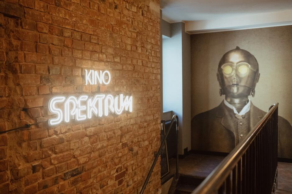 Na zdjęciu widoczny jest neonowy napis Kino Spektrum  umieszczony na ceglanej ścianie, tworzący ciekawy, industrialny klimat. Obok napisu, na końcu korytarza, znajduje się duży mural przedstawiający postać w masce gazowej, co dodaje wnętrzu nieco tajemniczego charakteru