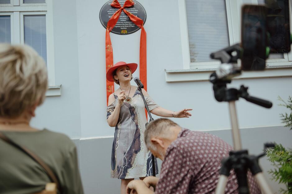 Na zdjęciu widzimy kobietę w czerwonym kapeluszu i kolorowej sukience, śpiewającą do mikrofonu podczas uroczystości odsłonięcia tablicy pamiątkowej. W tle znajduje się tablica ozdobiona czerwoną wstęgą, a na pierwszym planie widać publiczność i statyw z kamerą