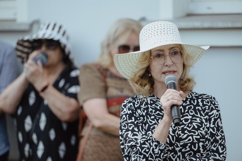 Na zdjęciu widzimy kobietę w białym kapeluszu i okularach, przemawiającą do mikrofonu. W tle stoją dwie inne kobiety, jedna z nich w czarno-białym kapeluszu, trzymająca napój, a druga w okularach przeciwsłonecznych