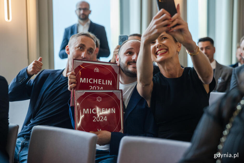 Na zdjęciu widać troje osób robiących sobie selfie, z czego dwóch mężczyzn trzyma certyfikaty Michelin 2024, a kobieta po prawej stronie uśmiecha się szeroko do telefonu (robi selfie). W tle siedzą inni uczestnicy wydarzenia, a jeden mężczyzna stoi i patrzy w kierunku aparatu.