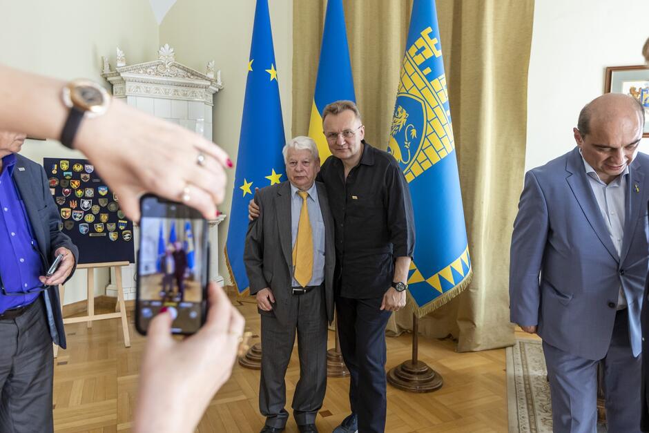 Trzej mężczyźni na tle statywów z flagami Unii Europejskiej, Ukrainy i Lwowa