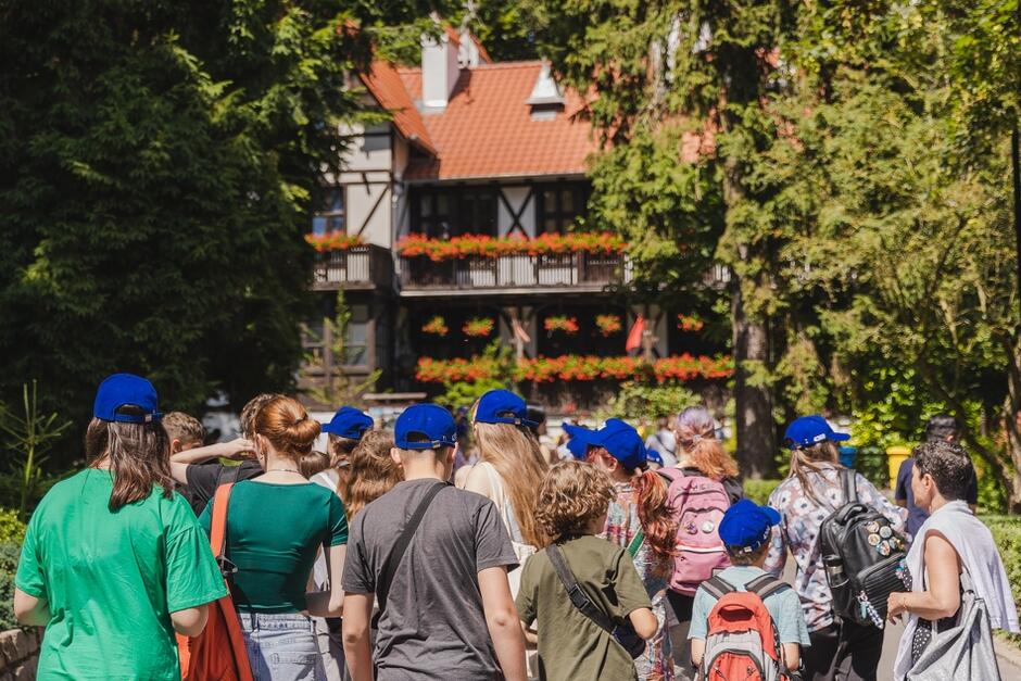 Na zdjęciu widać grupę ludzi, w większości dzieci i młodzieży, noszących niebieskie czapki, idących w kierunku budynku z czerwonym dachem i balkonami ozdobionymi kwiatami. W tle widoczne są drzewa, które otaczają budynek 