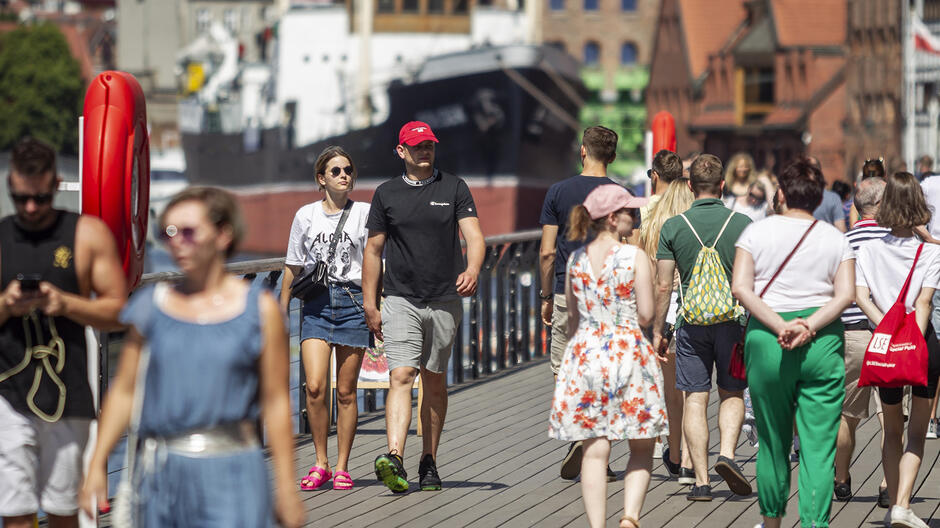 Na zdjęciu widać tłum ludzi spacerujących po promenadzie w słoneczny dzień. W tle znajdują się budynki oraz zacumowany statek