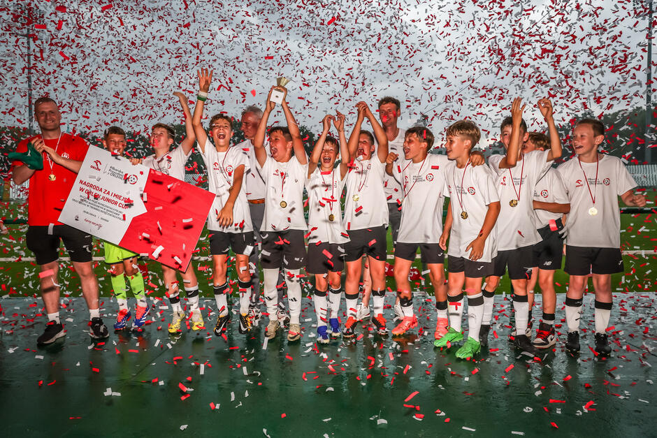 Na zdjęciu widać młodą drużynę piłkarską świętującą zwycięstwo, otoczoną deszczem konfetti. Zawodnicy w białych koszulkach i czarnych spodenkach trzymają puchar i duży czek, ciesząc się ze zdobytego trofeum i pierwszego miejsca w turnieju.