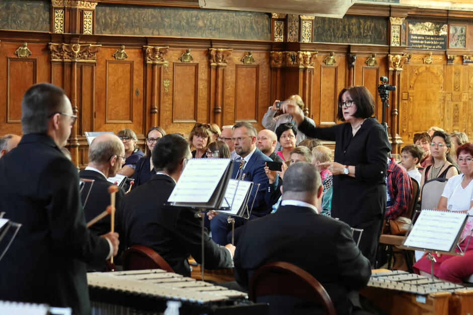 Na zdjęciu znajduje się orkiestra podczas występu w zabytkowej sali z drewnianymi zdobieniami na ścianach. Dyrygentka kieruje muzykami, a publiczność uważnie obserwuje koncert