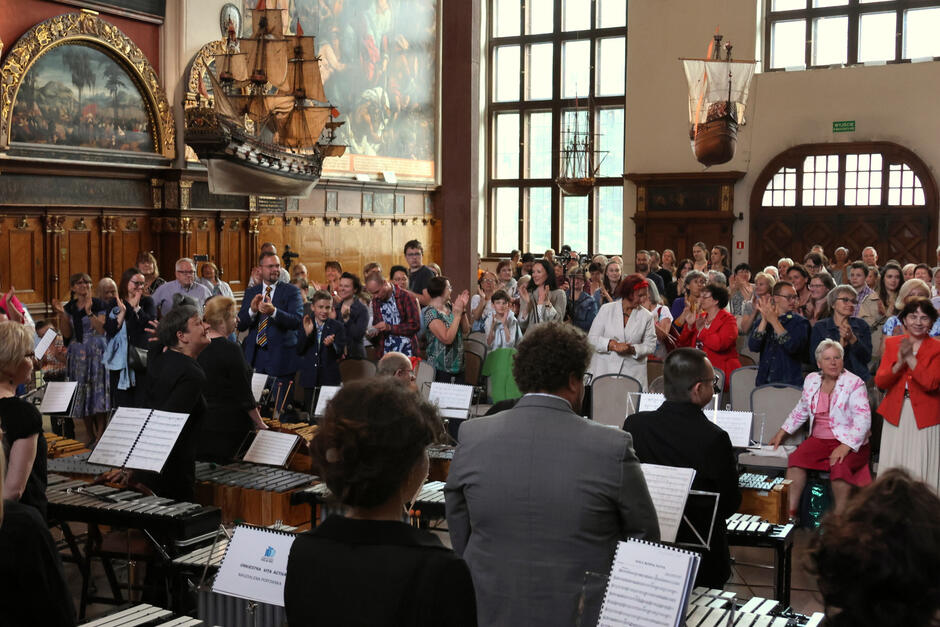 Na zdjęciu widać publiczność stojącą i oklaskującą orkiestrę po zakończeniu występu w pięknie zdobionej sali. W tle można dostrzec model statku wiszący pod sufitem oraz duże, ozdobne obrazy i okna