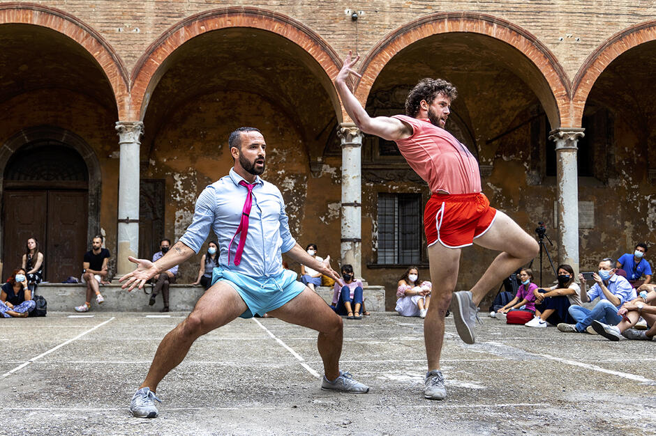 Zdjęcie przedstawia dwóch mężczyzn w kolorowych, sportowych strojach wykonujących energiczny taniec na dziedzińcu zabytkowego budynku. W tle siedzi publiczność, obserwując ich występ, a niektórzy z widzów mają na sobie maseczki ochronne