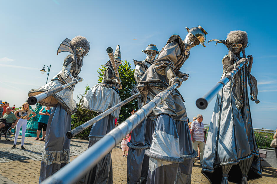 Zdjęcie przedstawia artystów z Teatru Akt ubranych w srebrne, futurystyczne kostiumy podczas ulicznego parady. Wykonawcy, poruszający się na szczudłach i trzymający długie kije, przyciągają uwagę przechodniów i widzów zgromadzonych na deptaku w słoneczny dzień