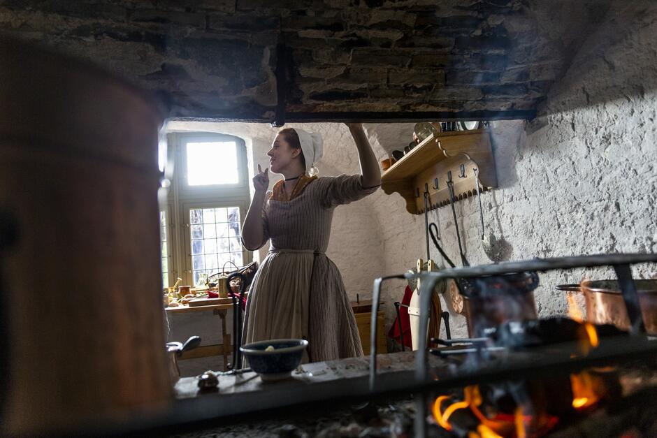 Na zdjęciu widać kobietę w stroju z epoki stojącą w historycznej kuchni. Otoczona jest starodawnymi naczyniami i narzędziami kuchennymi, a w tle widać ogień płonący w kominku.