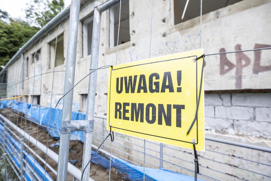 Zdjęcie przedstawia budynek w trakcie remontu, otoczony metalowym ogrodzeniem. Na ogrodzeniu widnieje żółta tablica z napisem UWAGA! REMONT , ostrzegająca przechodniów o trwających pracach.