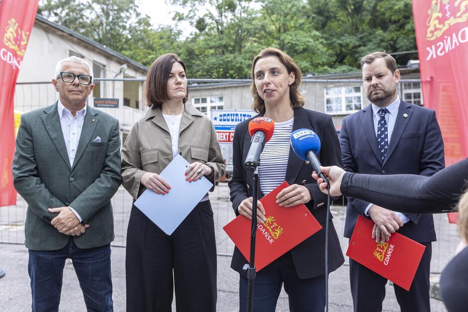 Zdjęcie przedstawia konferencję prasową zorganizowaną przed remontowanym budynkiem, w której uczestniczą cztery osoby: dwóch mężczyzn i dwie kobiety. Jedna z kobiet, trzymając czerwoną teczkę z herbem Gdańska, przemawia do mikrofonów oznaczonych logo lokalnych mediów.