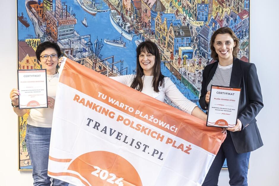 Na zdjęciu widzimy trzy uśmiechnięte kobiety trzymające certyfikaty i flagę z napisem Ranking Polskich Plaż 2024 - Travelist.pl . W tle znajduje się kolorowy obraz przedstawiający port lub nadmorskie miasto