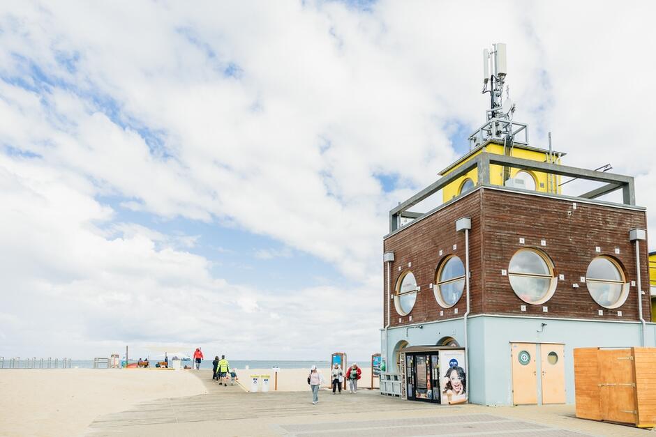 Zdjęcie przedstawia budynek znajdujący się przy plaży w Gdańsku, który ma okrągłe okna i jest wyposażony w antenę na dachu. Przed budynkiem znajdują się ludzie spacerujący po chodniku, a w tle widoczna jest plaża i morze pod częściowo zachmurzonym niebem.