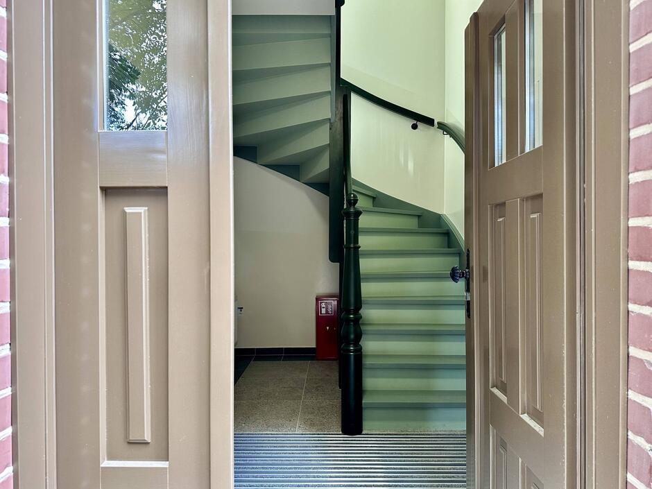 na zdjęciu uchylone drzwi wejściowe, widać wewnątrz budynku klatkę schodową i zielone schody drewniane