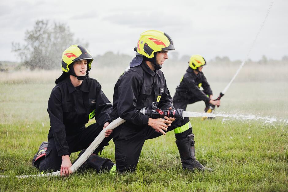 na zdjęciu dwóch strażaków, klękają na jedno kolano, trzymają węża strażackiego z którego leci strumień wody