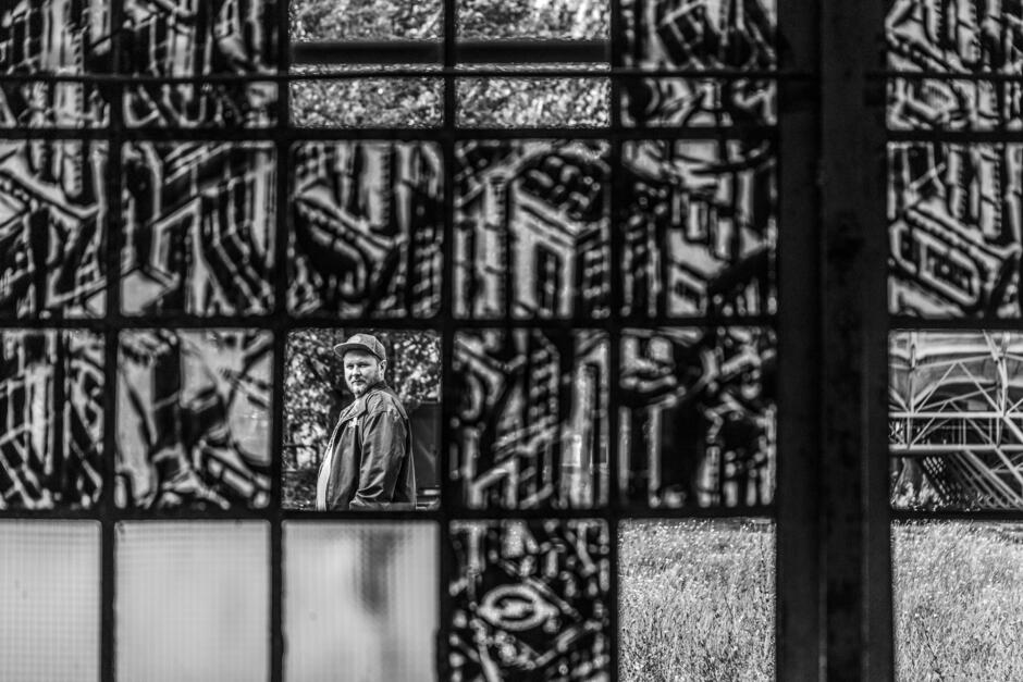 Na zdjęciu widzimy mężczyznę w czapce z daszkiem, który patrzy w stronę aparatu, stojąc na zewnątrz. Jego postać jest uchwycona przez ozdobne, rzeźbione okno, co tworzy efekt ramy wokół jego sylwetki, a całość jest wykonana w czarno-białej tonacji.