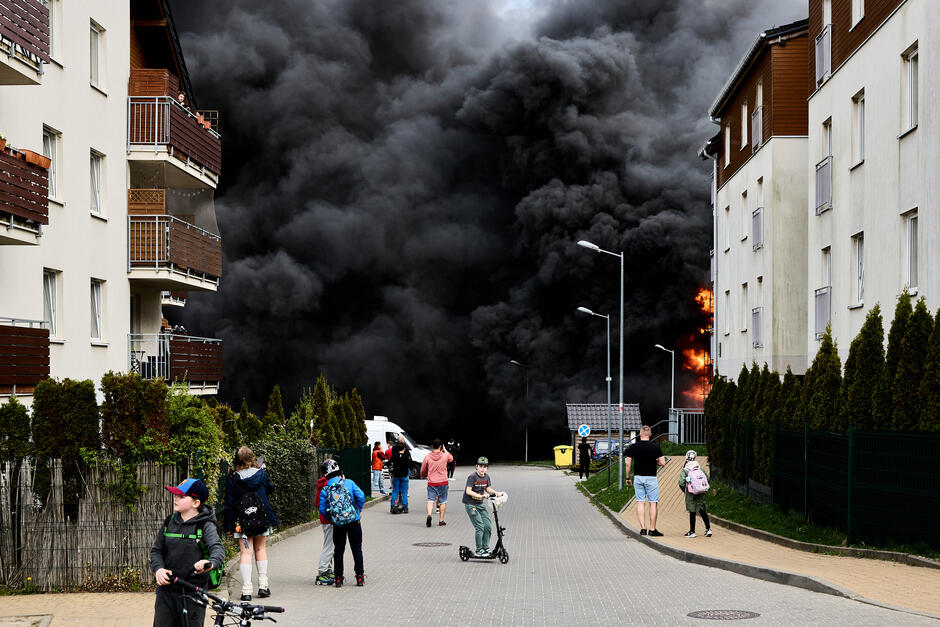 Na zdjęciu widzimy ulicę w osiedlu mieszkaniowym, po której spacerują ludzie, w tym dzieci na hulajnogach i rowerach. W tle unosi się gęsty, czarny dym z pożaru, który wybuchł w jednym z budynków, a płomienie są widoczne po prawej stronie obrazu.