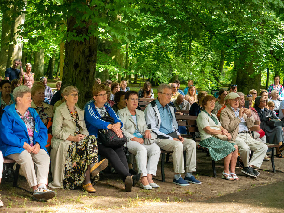 Na zdjęciu widzimy grupę starszych ludzi siedzących na ławkach w parku, otoczonych przez bujną zieleń drzew. Wydarzenie odbywa się na świeżym powietrzu, a uczestnicy wyglądają na zaangażowanych i skupionych na tym, co się dzieje przed nimi.