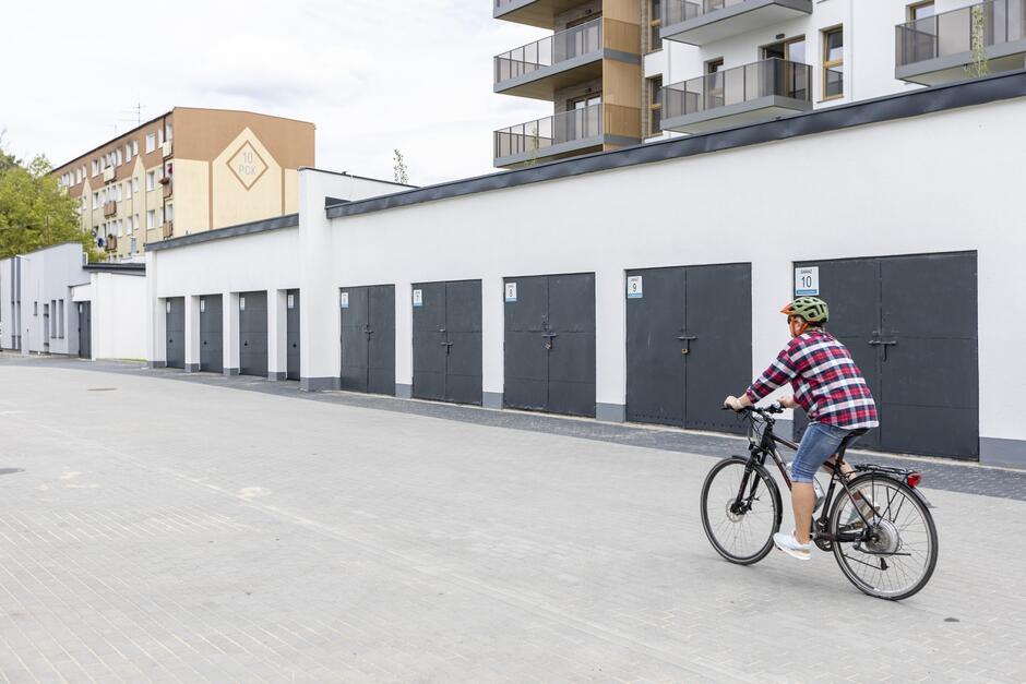 na zdjęciu chłopak jedzie na rowerze, po prawej stronie stoją garaże o białych ścianach i czarnych drzwiach