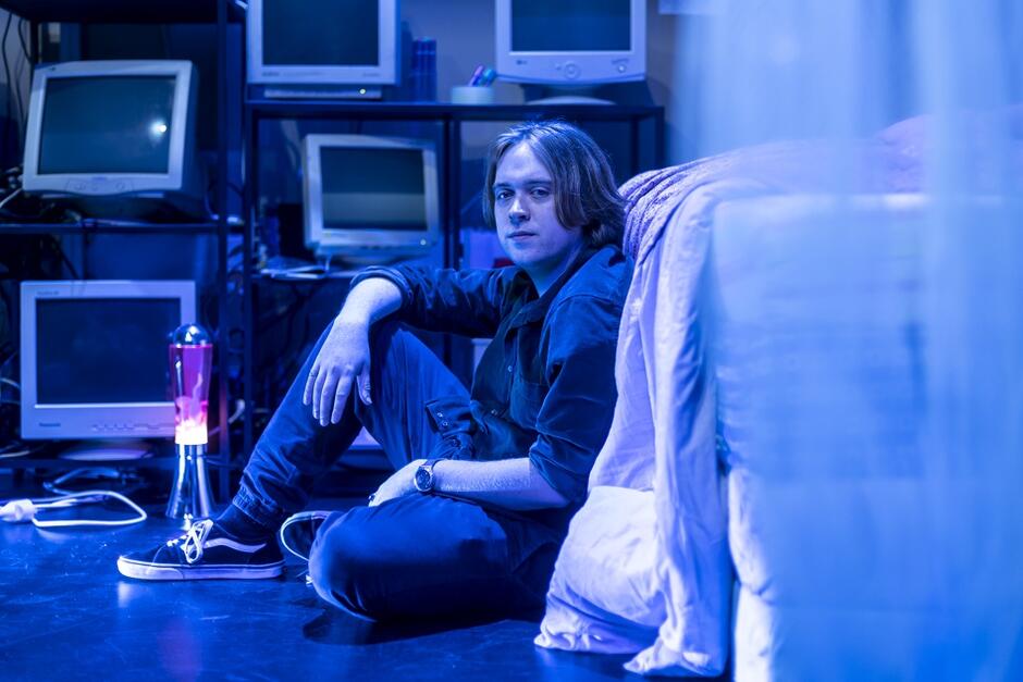 Zdjęcie przedstawia młodego mężczyznę siedzącego na podłodze obok łóżka, który patrzy w stronę aparatu. W tle znajduje się kilka starych monitorów, a cała scena jest oświetlona niebieskim światłem, co nadaje jej technologiczny i nostalgiczny klimat
