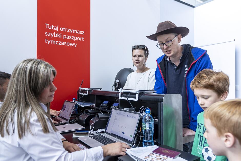 Na zdjęciu znajduje się stanowisko obsługi paszportowej, gdzie urzędniczka pracuje przy laptopie, obsługując klienta. Po drugiej stronie lady stoi mężczyzna w kapeluszu, kobieta oraz dwójka dzieci, którzy czekają na wydanie szybkiego paszportu tymczasowego, co jest oznaczone na czerwonym tle napisem Tutaj otrzymasz szybki paszport tymczasowy .