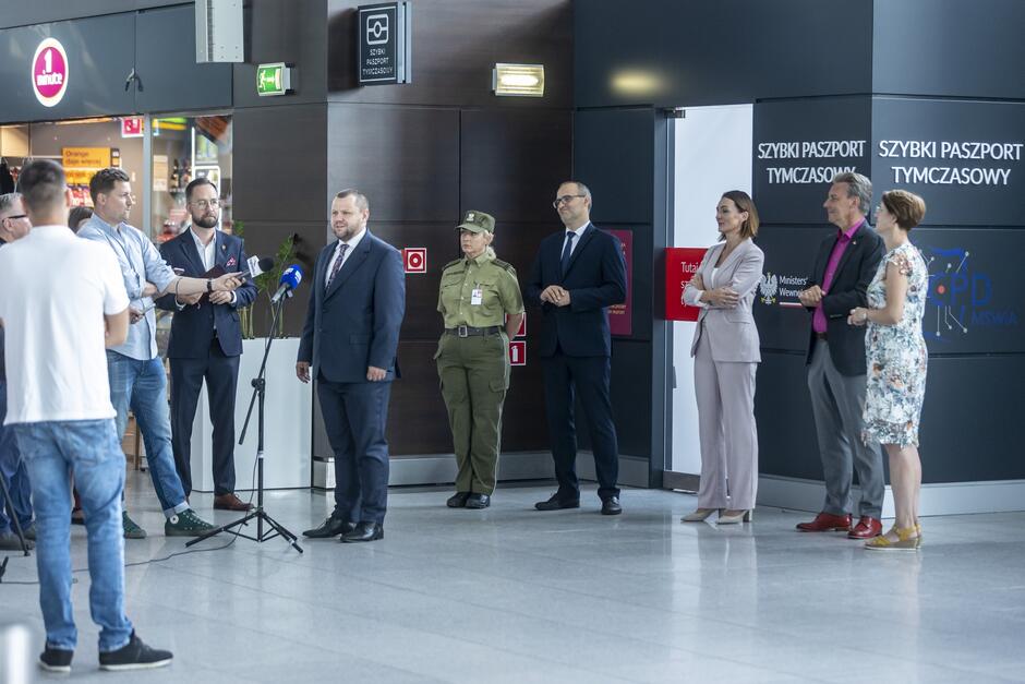 Na zdjęciu widzimy grupę ludzi stojących na lotnisku przed punktem oznaczonym jako Szybki Paszport Tymczasowy . W centralnym miejscu znajduje się mężczyzna w garniturze, przemawiający do mikrofonu, a obok niego stoją inni urzędnicy, w tym kobieta w mundurze służb granicznych.