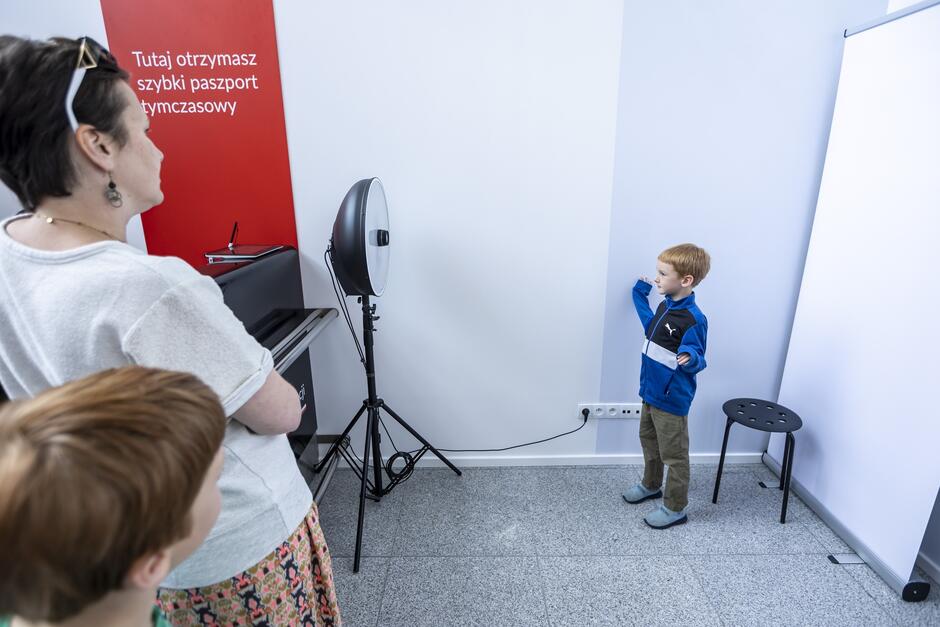 Na zdjęciu znajduje się mały chłopiec ubrany w niebieską kurtkę, stojący przed białym tłem, gotowy do zrobienia zdjęcia paszportowego. Obok niego stoi kobieta, prawdopodobnie jego matka, obserwując proces fotografowania, a na ścianie widnieje czerwony napis Tutaj otrzymasz szybki paszport tymczasowy .