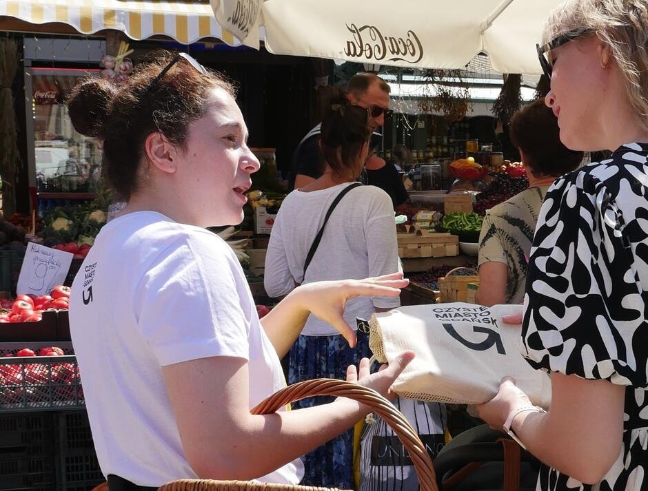 Na zdjęciu widzimy dwie kobiety rozmawiające na targu, jedna z nich trzyma wiklinowy kosz, a druga płócienną torbę z napisem CZYSTE MIASTO GDAŃSK . W tle widoczne są stragany z owocami i warzywami oraz kilku innych klientów robiących zakupy.