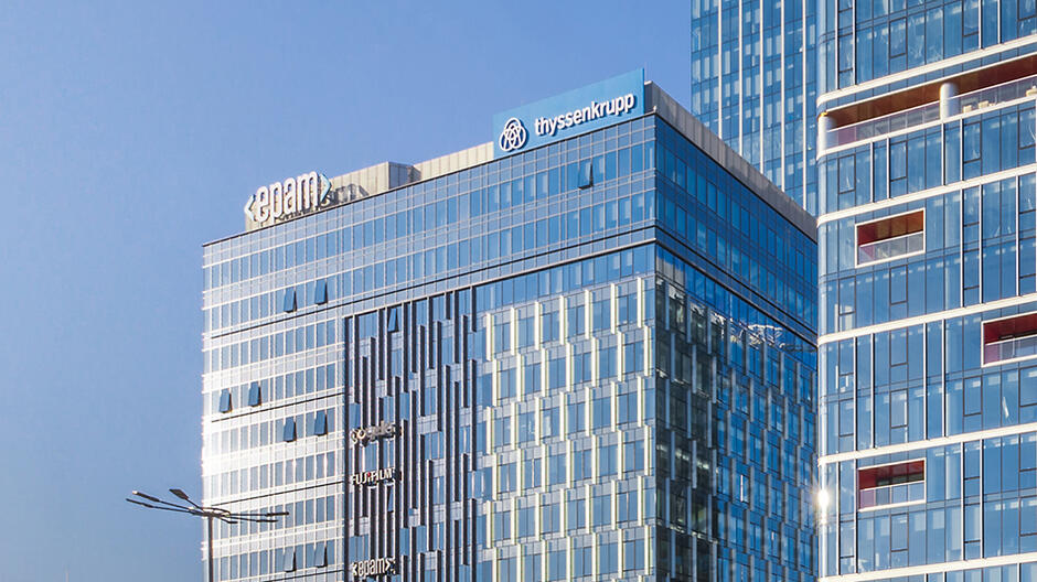 Na zdjęciu widzimy nowoczesny, szklany wieżowiec z logo firm EPAM i thyssenkrupp na jego szczycie. W tle widać inne wysokie budynki o podobnej, szklanej fasadzie, co nadaje scenerii biznesowy charakter.