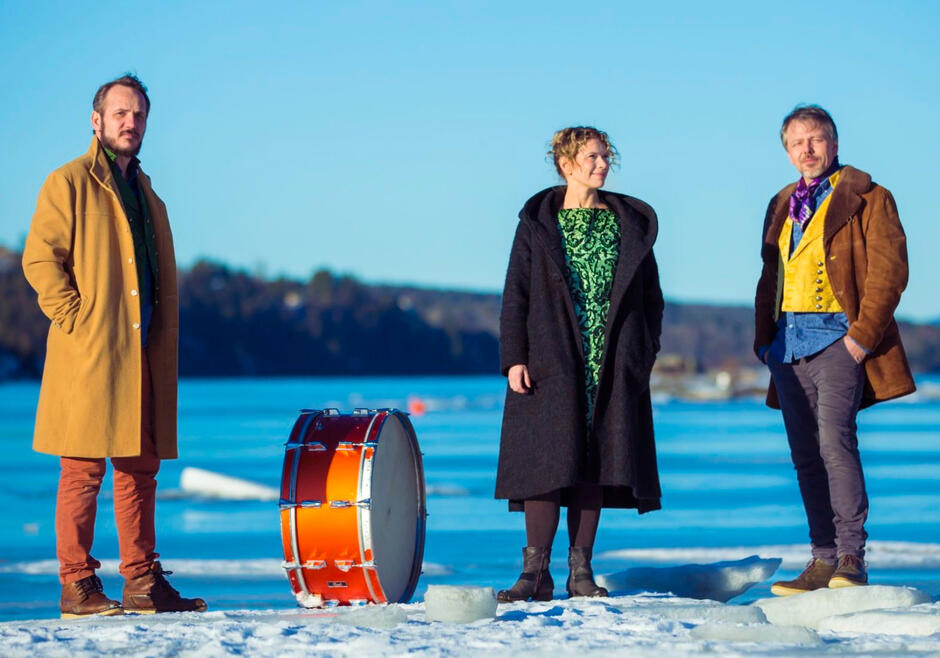 Na zdjęciu znajdują się trzy osoby stojące na zamarzniętym jeziorze, w tle widoczny jest zalesiony brzeg. Każda osoba ma na sobie zimowy płaszcz, a obok nich znajduje się duży, pomarańczowy bęben
