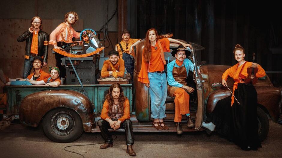 Na zdjęciu widzimy grupę jedenastu osób w pomarańczowych i brązowych ubraniach, które pozują wokół starej ciężarówki w warsztacie lub garażu. Jedna z osób trzyma skrzypce, podczas gdy inne stoją, siedzą lub opierają się o ciężarówkę