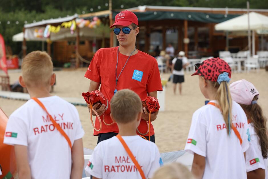 Na zdjęciu młody ratownik w czerwonej koszulce i czapce, prowadzi zajęcia na plaży dla grupy dzieci, które noszą białe koszulki z napisem Mały Ratownik . W tle widać plażowe zaplecze z ludźmi oraz różne dekoracje.