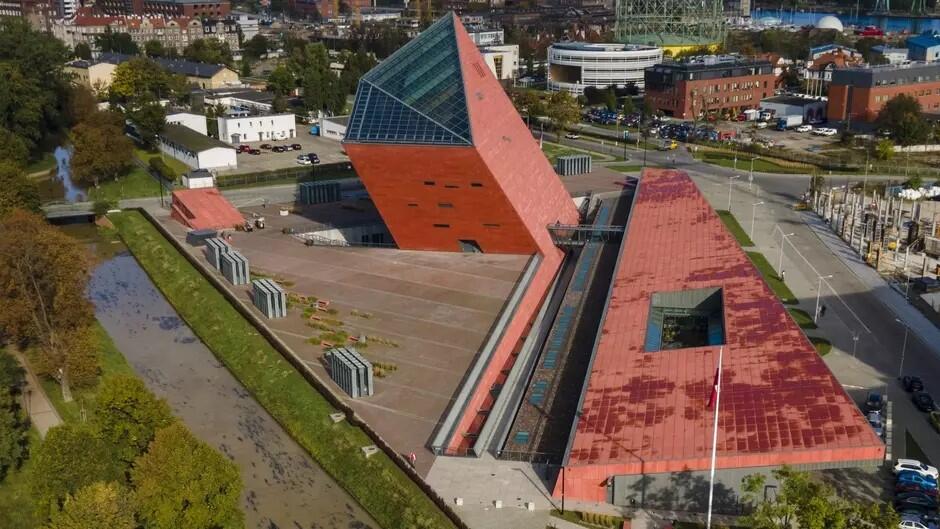 Fragment przestrzeni miejskiej, w centrum na pierwszym planie nowoczesny budynki - z lewej strony wyższa część pokryta czerwoną elewacją, z prawej niższa  