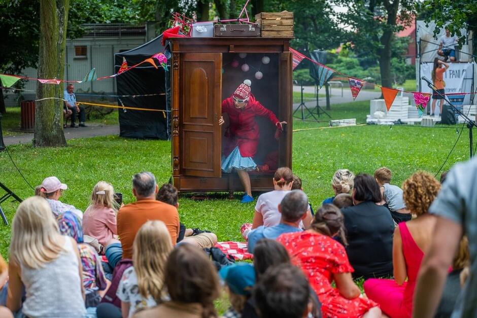 Zdjęcie przedstawia plenerowy spektakl teatralny, w którym aktor wychodzi z dużej, drewnianej szafy, ubrany w czerwony płaszcz i okulary przeciwsłoneczne. Widzowie, siedzący na trawie przed sceną, obserwują występ, który odbywa się w otoczeniu drzew i kolorowych girland
