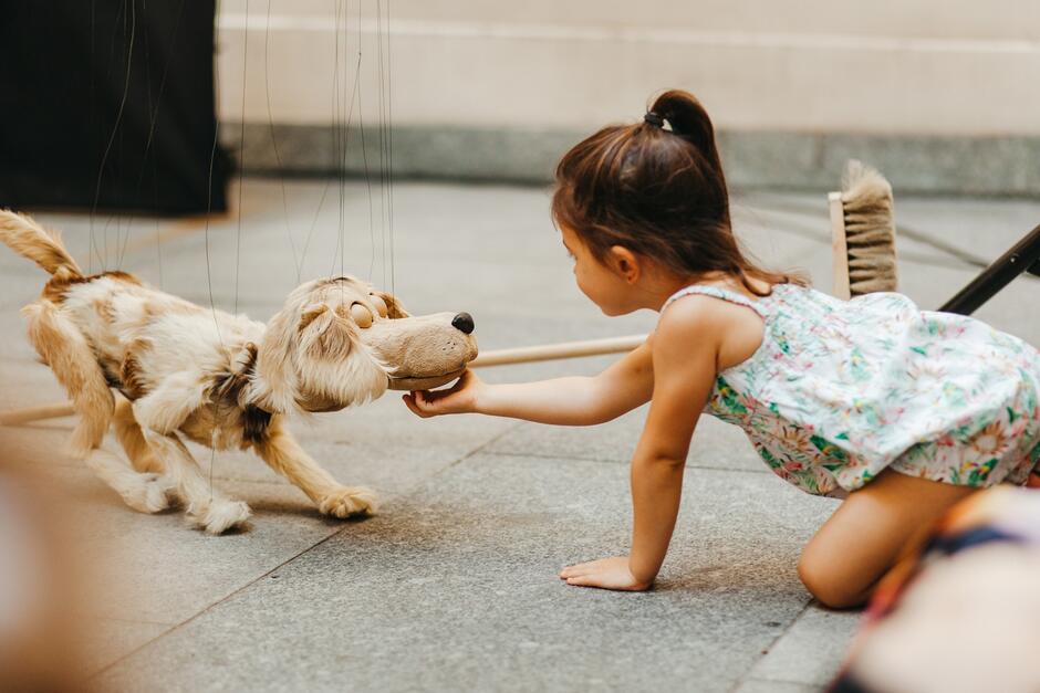 Zdjęcie przedstawia małą dziewczynkę w jasnej, kwiecistej sukience, bawiącą się z marionetkowym psem. Dziewczynka klęczy na podłodze i delikatnie dotyka nosa marionetki, podczas gdy piesek jest manipulowany za pomocą sznurków