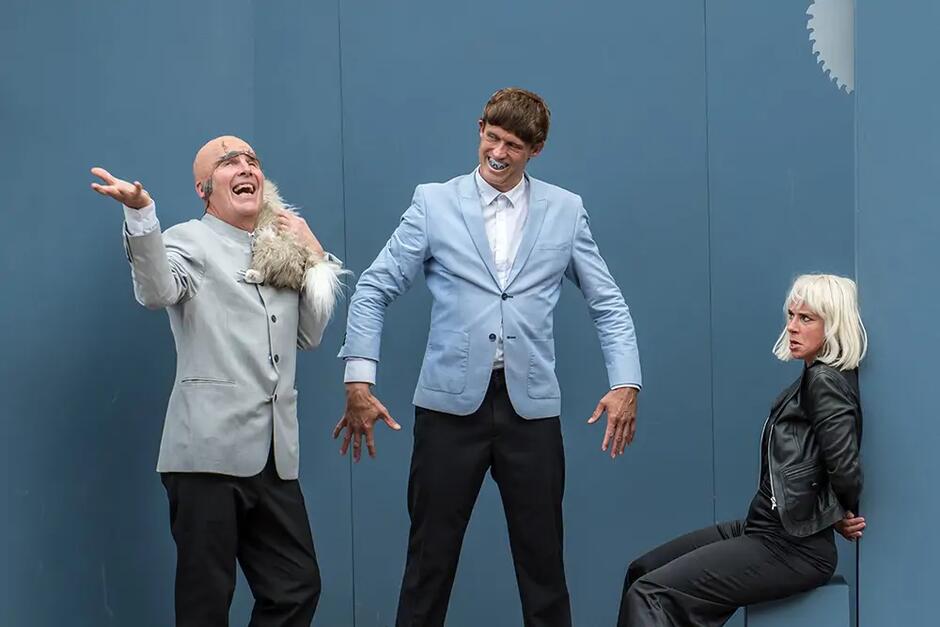 Zdjęcie przedstawia trójkę aktorów podczas występu teatralnego. Dwóch mężczyzn w jasnych marynarkach odgrywa komiczne role, podczas gdy kobieta w czarnej kurtce siedzi z boku z wyrazem zaskoczenia na twarzy