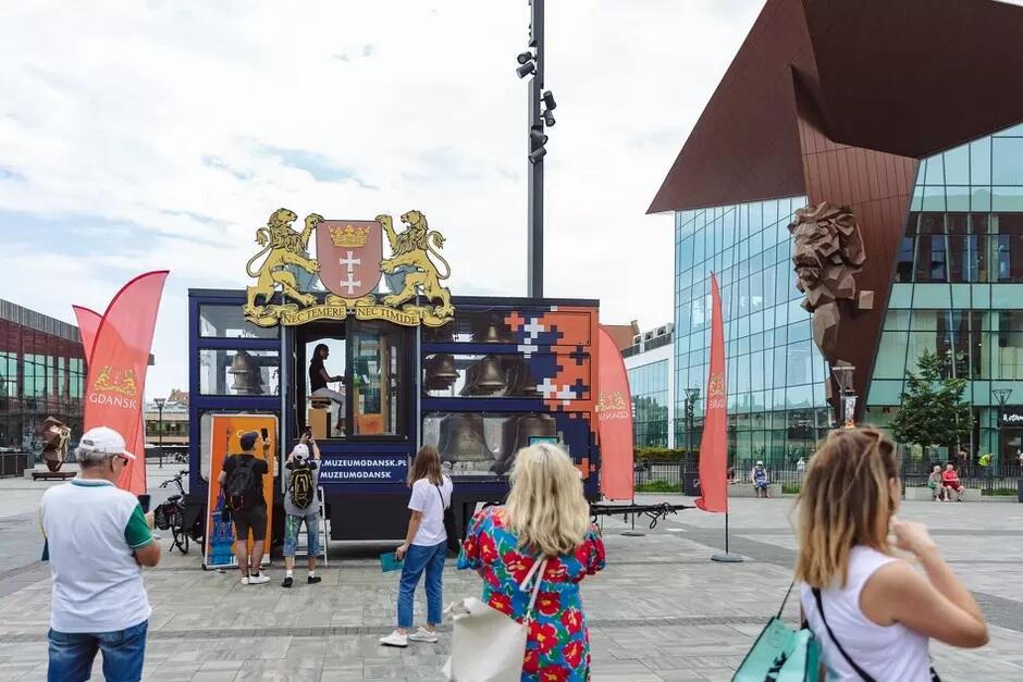 Zdjęcie przedstawia mobilny carillon Muzeum Gdańska na placu z grupą ludzi, którzy go oglądają. W tle widoczne są nowoczesne budynki oraz charakterystyczna rzeźba lwa, a całość ozdobiona jest flagami z logo Gdańska