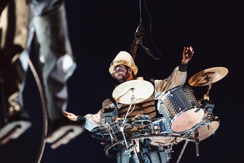 Zdjęcie przedstawia artystę bębniarza zawieszonego w powietrzu na linie, grającego na zestawie perkusyjnym podczas występu nocą. Postać jest ubrana w kostium sceniczny i ma pomalowaną twarz, co dodaje widowisku teatralnego charakteru