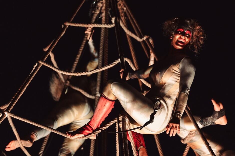 Zdjęcie przedstawia artystkę ubraną w obcisły kostium i czerwone buty, wykonującą akrobacje na linach podczas nocnego przedstawienia. Postać ma pomalowaną twarz i wygląda na roześmianą, co dodaje dynamiki i ekspresji całej scenie