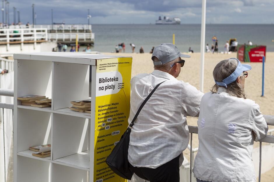 Na zdjęciu widać plażę z biblioteczką na plaży, oferującą książki do wypożyczenia na świeżym powietrzu. Na pierwszym planie znajduje się para starszych ludzi, patrzących na morze, a w tle widoczny jest statek na horyzoncie.