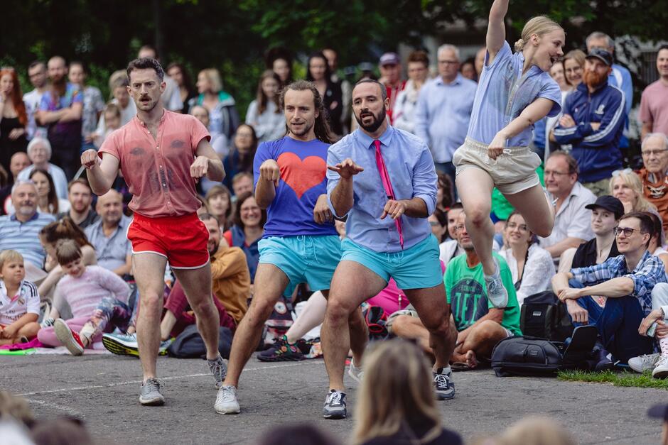 Na zdjęciu widać czterech młodych tancerzy wykonujących dynamiczny występ na świeżym powietrzu, przed licznie zgromadzoną publicznością. Ubrani w kolorowe stroje, tancerze wykonują synchronizowane ruchy, a jedna z tancerek jest w trakcie skoku.