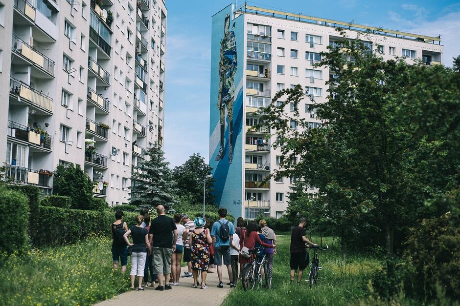 Zdjęcie przedstawia grupę ludzi stojących na chodniku pomiędzy dwoma wysokimi blokami mieszkalnymi, oglądających duży mural na ścianie jednego z budynków. Mural przedstawia postać człowieka, a w tle widać otoczenie zielenią oraz balkonami pełnymi roślinności.