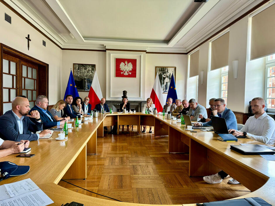 Kilkanaście osób siedzi wokół stołów ustawionych w obwarzanek w sali urzędu i debatuje. W tle godło państwowe oraz flagi - polskie i unijne. 