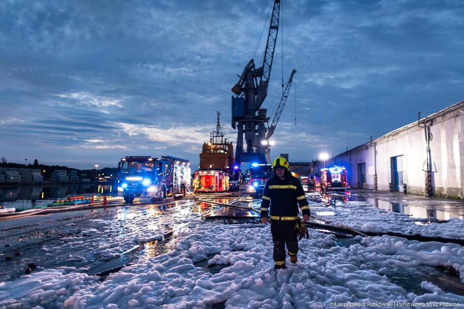 Zdjęcie z akcji gaśniczej. Strażak idzie po nabrzeżu portowym, które jest pokryte białą pianą gaśniczą, co wygląda jakby spadł śnieg