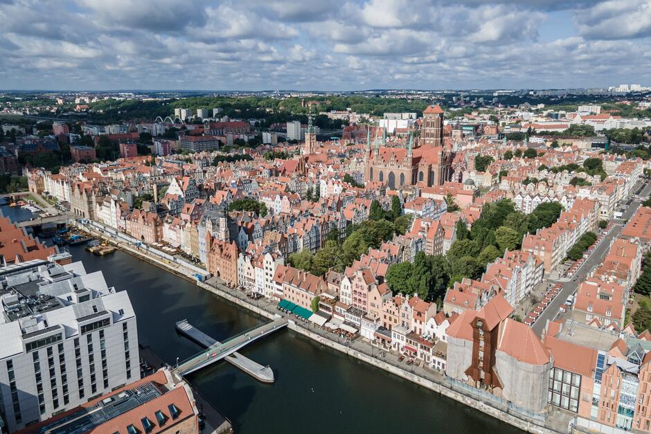 Zdjęcie z lotu ptaka przedstawiające gdańska starówkę. Na pierwszym planie rzeka Motława, dalej budynki Głównego Miasta z kamienicami i wieżami ratusza oraz kościoła.