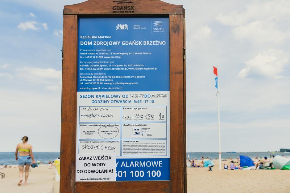 Tablica informacyjna na plaży. Na tablicy kartka informująca o zakazie kąpieli z powodu skażenia wody. W tle ludzie na plaży.