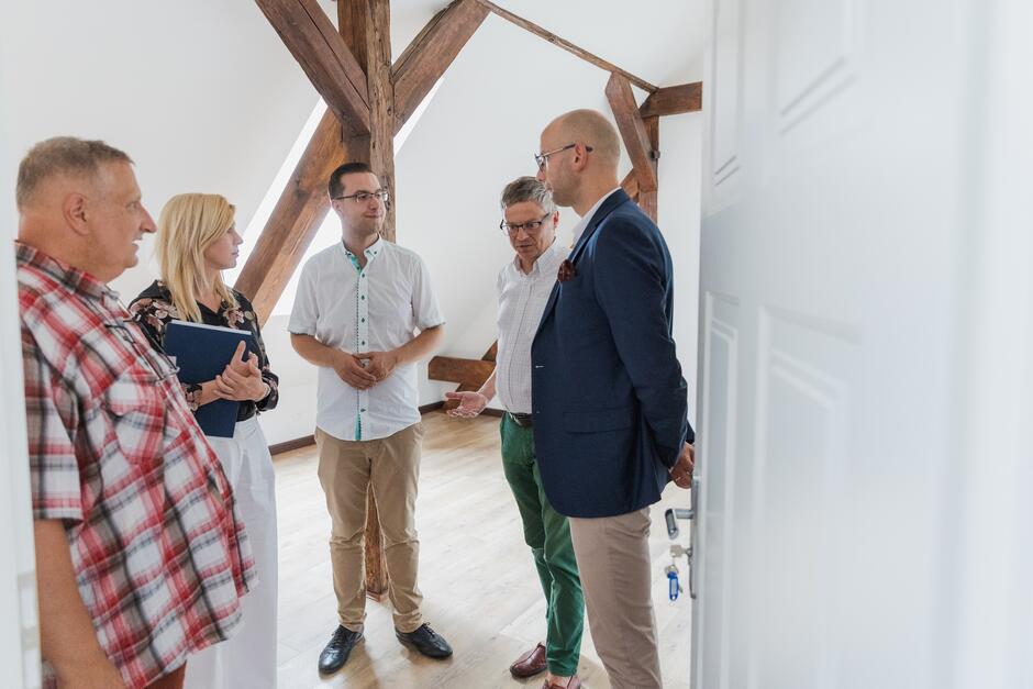 Na zdjęciu widzimy pięć osób znajdujących się w jasnym pomieszczeniu z drewnianymi belkami stropowymi, co sugeruje, że jest to poddasze lub wyższe piętro budynku. Grupa ludzi wygląda, jakby brała udział w spotkaniu lub dyskusji.