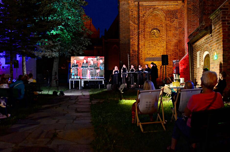 Zdjęcie przedstawia nocny koncert na świeżym powietrzu, odbywający się przy zabytkowym budynku z cegły. Widownia siedzi na leżakach i ławkach, oglądając występ na żywo, który jest również wyświetlany na dużym ekranie, a scena i drzewa w tle są kolorowo oświetlone.