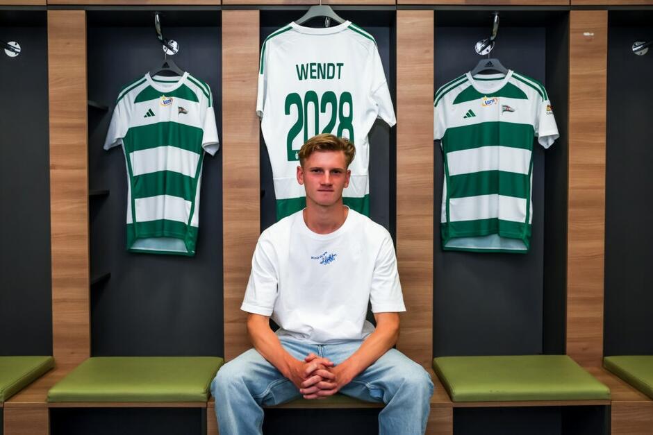 Młody blond mężczyzna pozuje do zdjęcia, siedząc w piłkarskiej szatni. Za nim wiszą biało-zielone koszulki